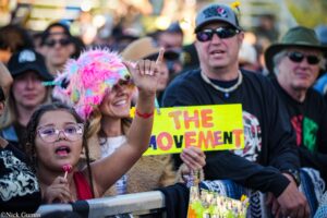 Young girl with braided hair and pink glasses holds a pink lollipop toward the camera as a crowded protest scene unfolds, a raised hand and a bright yellow sign with red letters in the foreground.