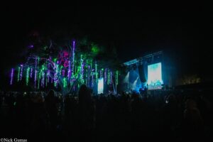 Night concert with trees wrapped in purple and green lights; stage with a large screen on the right and a silhouetted crowd.