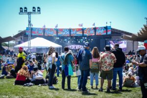 Crowd at an outdoor music festival facing a colorful stage banner that reads 'Cali Roots' with bright lights overhead.