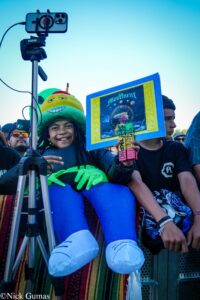 Smiling child in a bright emoji hat holds a framed poster that says 'The Movement' at an outdoor event.