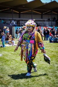 Colorful festival performer wearing psychedelic outfit and sunglasses, walking on grass at an outdoor event with people in the background.