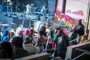 Crowd of people at a live outdoor concert, a colorful banner waving in the foreground with the stage and musicians in the background.