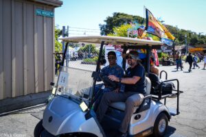 Two men ride in a gray Yamaha golf cart at a fairground area, with a colorful flag attached to the cart and a sign reading 'PAUL VIEReGGE WAY' on a wooden building to the left; background shows booths and people walking.