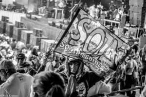 Crowd of people at a street festival, a person holds a large graffiti-style banner above the crowd.