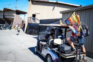 Group of people standing and walking near a beige industrial building as a blue golf cart with a colorful flag passes by in the foreground on a sunny day, blue sky overhead