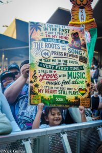 Colorful poster with lyrics and tropical designs held up at a concert by cheering fans.