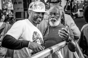 Two men lean over a railing at an outdoor event, sharing a smile as they chat; one wears a white shirt and a patterned cap, the other has gray hair and a gray beard.