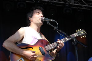 Male guitarist singing into a microphone on stage while playing a sunburst acoustic guitar with a strap over his shoulder