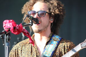 Musician with curly hair and sunglasses singing into a microphone on stage, wearing a patterned shirt and a blue guitar strap.