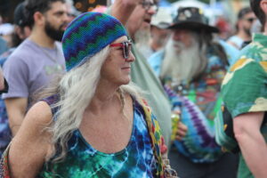 Woman in colorful tie-dye top and blue knit hat with green pattern, wearing red-framed sunglasses, in a crowded outdoor event.