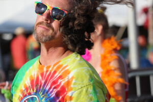 Man at a festival wearing rainbow sunglasses and a bright tie-dye shirt, shown in a close-up side profile.