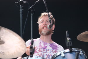Drummer with a beard singing into a microphone while playing drums, wearing a pink-purple tie-dye shirt.