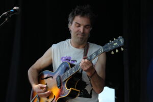 Male guitarist performing on a dark stage, playing a sunburst hollow-body guitar and looking down at the neck.