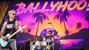 Blond guitarist in sunglasses plays a mint-green electric guitar on a colorful tropical stage; drummer in the back.