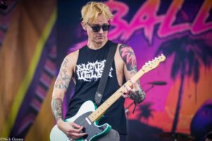 Male guitarist wearing sunglasses and a black sleeveless shirt, playing a turquoise electric guitar on a colorful stage backdrop.