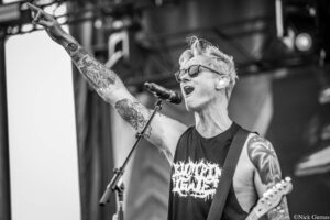 Rock musician singing into a microphone on stage, arm raised, tattooed forearm visible in grayscale photo.