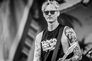 Man wearing sunglasses and a sleeveless shirt, playing an electric guitar with tattooed arms during a performance (black and white photo).