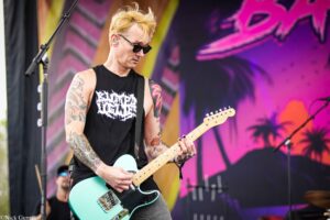 Guitarist with blond hair, sunglasses, and tattoos plays a turquoise electric guitar on a colorful stage backdrop.
