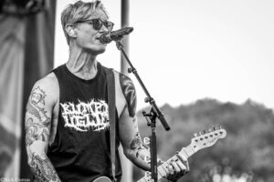 Black-and-white photo of a tattooed male guitarist singing into a microphone on stage, wearing sunglasses and a sleeveless shirt.