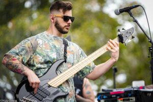 Male bassist in a camo shirt wearing sunglasses and in-ear monitors plays a black bass outdoors.