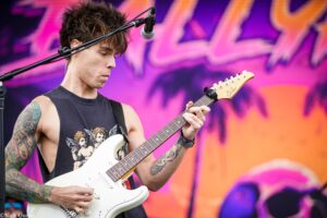 Male guitarist playing a white electric guitar on stage in a sleeveless shirt; colorful backdrop behind him with pinks and purples.