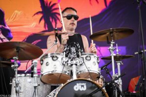 Drummer wearing sunglasses plays a silver DW drum kit on a colorful stage backdrop.