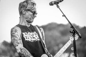 Guitarist with tattoos and glasses, playing a white electric guitar on an outdoor stage.