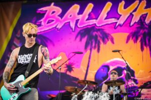 Guitarist with blond hair and sunglasses plays a turquoise electric guitar on a bright pink-orange stage backdrop.