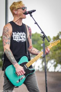 Male guitarist with blond hair and sunglasses singing into a microphone while playing a teal electric guitar outdoors, tattoos visible on arms.