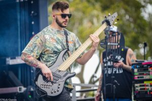 Musician wearing a camo shirt and sunglasses playing a gray bass guitar on an outdoor stage, with equipment nearby.