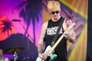 Guitarist playing a turquoise electric guitar on a colorful stage backdrop, wearing a black sleeveless shirt and sunglasses.