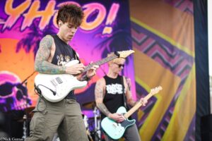 Guitarist with tattoos playing a white electric guitar on a colorful stage backdrop at a live concert.