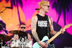 Guitarist with blond hair and tattoos plays a mint-green electric guitar on a vibrant pink stage backdrop.