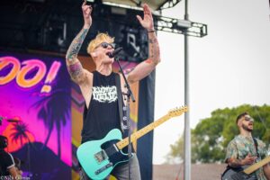 Rock musician with teal electric guitar performs on stage, tattoos visible on arms, singing into a microphone with hands raised.