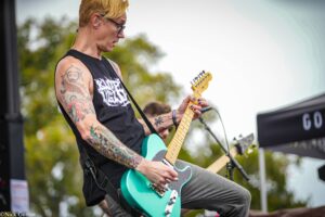 Guitarist with blond hair and tattoos playing a turquoise electric guitar on an outdoor stage, wearing a black sleeveless shirt.