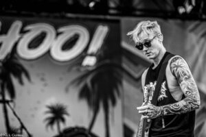 Guitarist with tattoos and sunglasses performing on stage, concentrating on his guitar in a black-and-white photo.
