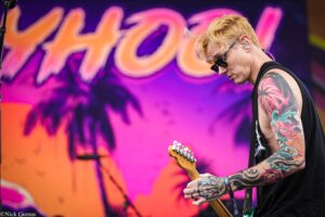Guitarist with colorful tattoos playing an electric guitar on a vibrant concert stage backdrop with pink and orange hues