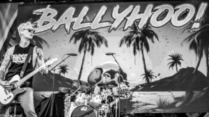 Black-and-white photo of a guitarist with sunglasses and tattoos playing an electric guitar on stage, with a drummer behind him and a tropical backdrop.