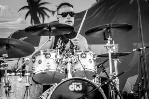 Black-and-white photo of a drummer wearing sunglasses, playing a DW drum kit on stage during a live performance.