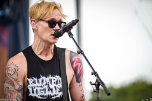 Male guitarist with blond hair and sunglasses singing into a microphone at an outdoor show, wearing a black sleeveless shirt and displaying colorful tattoos on both arms.