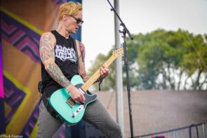 Male guitarist with tattoos playing a turquoise electric guitar on an outdoor stage wearing black sleeveless shirt and sunglasses.