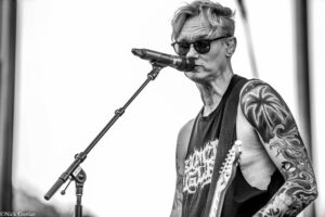 Male guitarist wearing sunglasses and a sleeveless shirt sings into a microphone on stage, displaying tattooed arms.