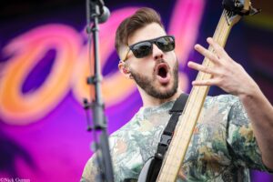 Bass guitarist wearing sunglasses and a camo shirt plays a bass on a vibrant stage backdrop of pink and purple lights.