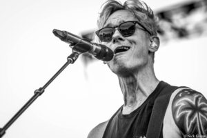 Male singer wearing sunglasses, singing into a microphone on stage with a guitar strap over his shoulder (black-and-white photo).