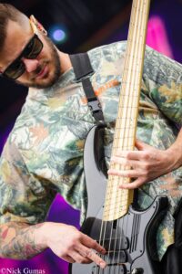 Musician in camouflage shirt wearing sunglasses plays a bass guitar on stage, close-up of hands on strings and neck.