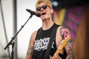Male guitarist wearing sunglasses, singing into a microphone on stage with a black tank top and colorful tattoos visible on his arms and shoulders.