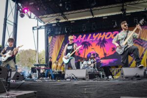 Live rock band performing on an outdoor stage with a vibrant tropical banner in the background; guitarist left, vocalist/guitarist center, and bassist right in sunglasses, drums at back.