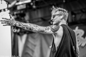 Man with full arm tattoo reaching toward the camera during a live performance, wearing sunglasses in black and white