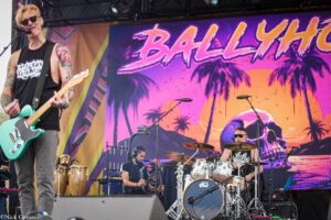 Male guitarist with turquoise electric guitar performing on stage, colorful tropical backdrop and drummer in the foreground.