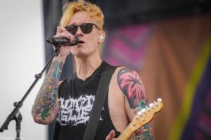 Male rock singer with blond hair and visible tattoos, wearing sunglasses and a black sleeveless shirt, singing into a microphone on stage